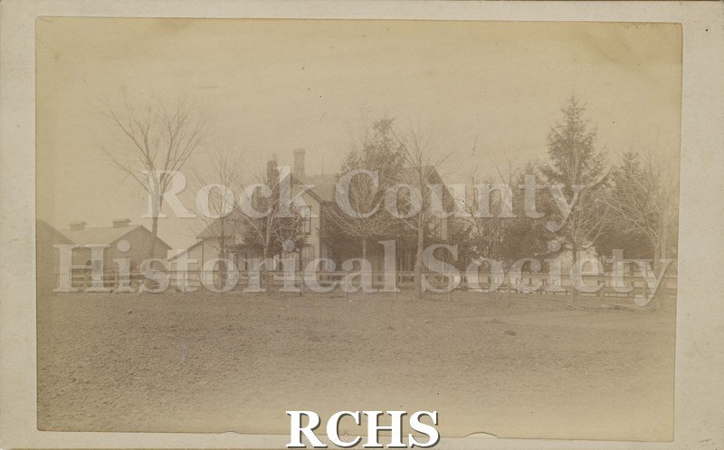 Black and white photograph of Rockwood's farmhouse, a two-story farmhouse with barns to the left; a wooden fence in front as well as trees behind the fence.
