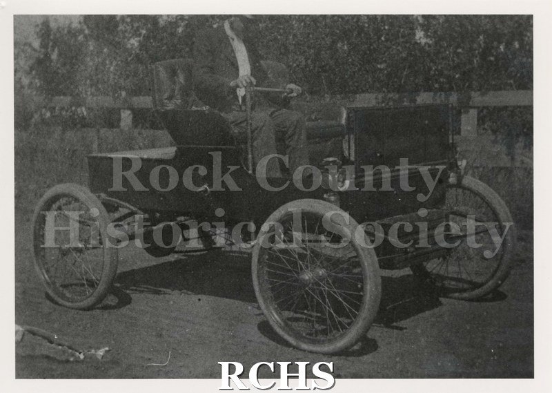 Black and white photograph of a man seated in an early automobile.