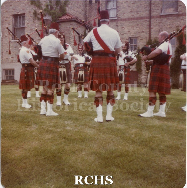 Colored photograph of of bagpipers performing on lawn outside Old Armory building, Janesville. Written on reverse: " Shriner's Parade, 1984".