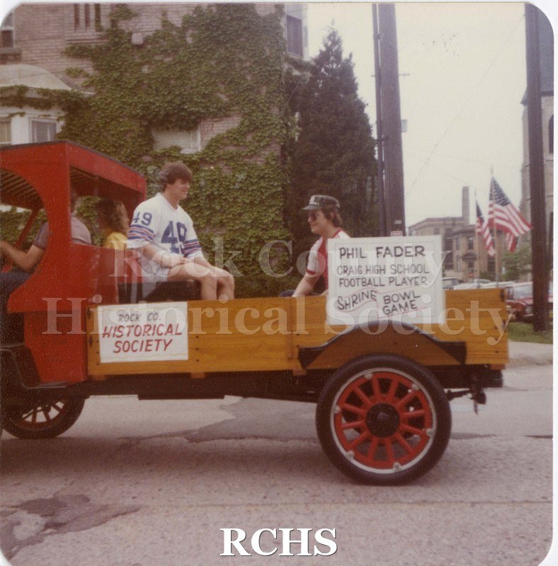 Colored photograph of of an antique motorized truck with two males riding in its bed. Signs on side of truck: "ROCK CO.