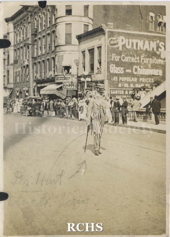 Black and white photograph of a parade on Milwaukee Street with "Uncle Sam". A man in an Uncle Sam costume is marching in parade on Milwaukee St., Janesville.