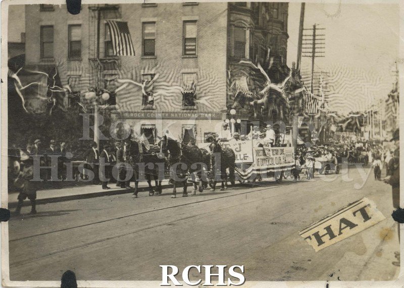 Black and white photograph of a July 4th parade ca 1917 with Rock County National Bank, In background Team of four horses pulling float over bridge.
