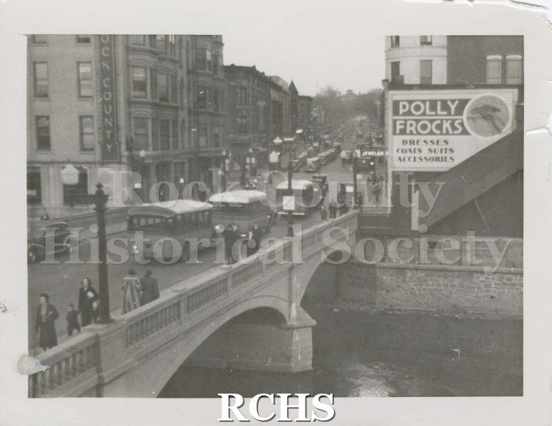 Black and white photograph of Main Street Janesville, WI. Shows bridge over Rock River, pedestrians strolling along sidewallk over bridge; and three buses on street.