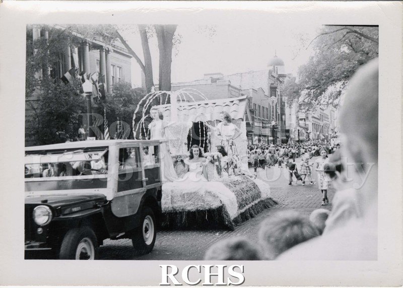 Black and white photograph of a parade float drawn by motorized vehicle. Float is carrying three young women in party dress.