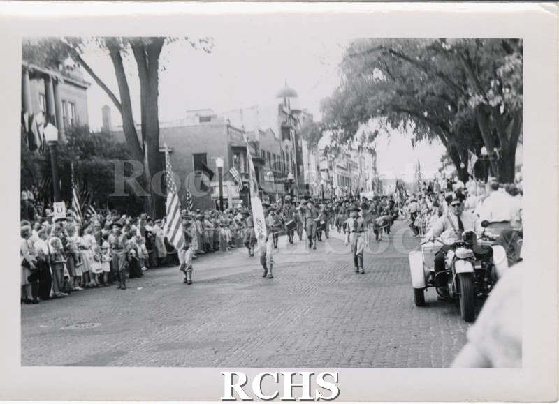 Black and white photograph of a parade marchers on South Main Street, Janesville, with public library and other downtown buildings in view.