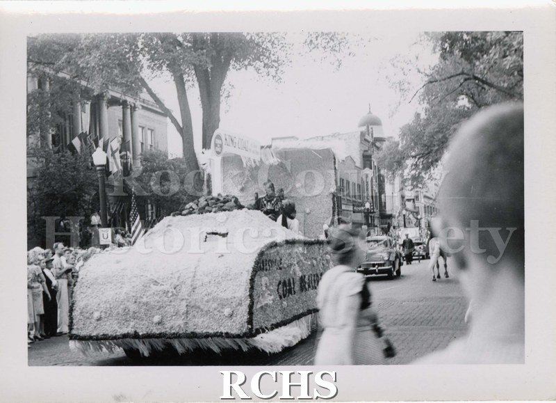 Black and white photograph of a parade float. Side of float advertises "Janesville Coal Dealer(?). Public library on South Main St. Janesville appears in background.