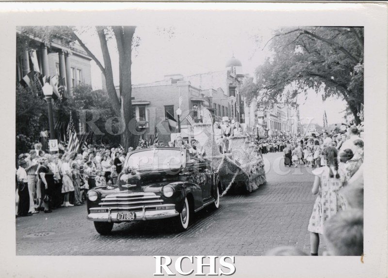 Black and white photograph of a parade float drawn by a period vehicle along South Main Street, Janesville, in front of public library. Written on reverse: "c. 1941".