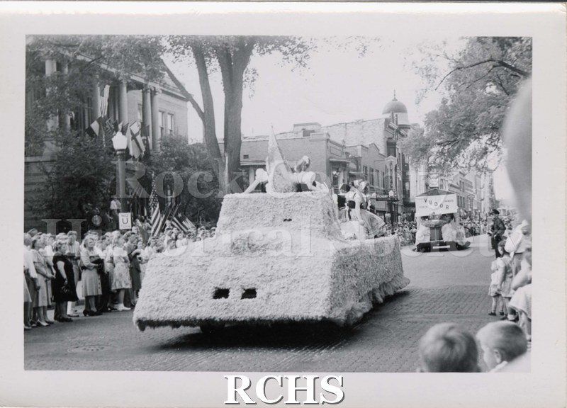 Black and white photograph of a parade floats on South Main Street, Janesville, in front of Carnegie Library. Written on reverse: "c. 1941".