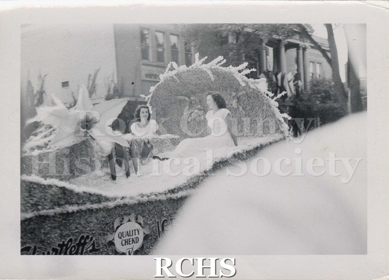 Black and white photograph of a parade float carrying two young women. "QUALITY BREAD" sign on side of float. Carnegie Library, Janesville, in background.