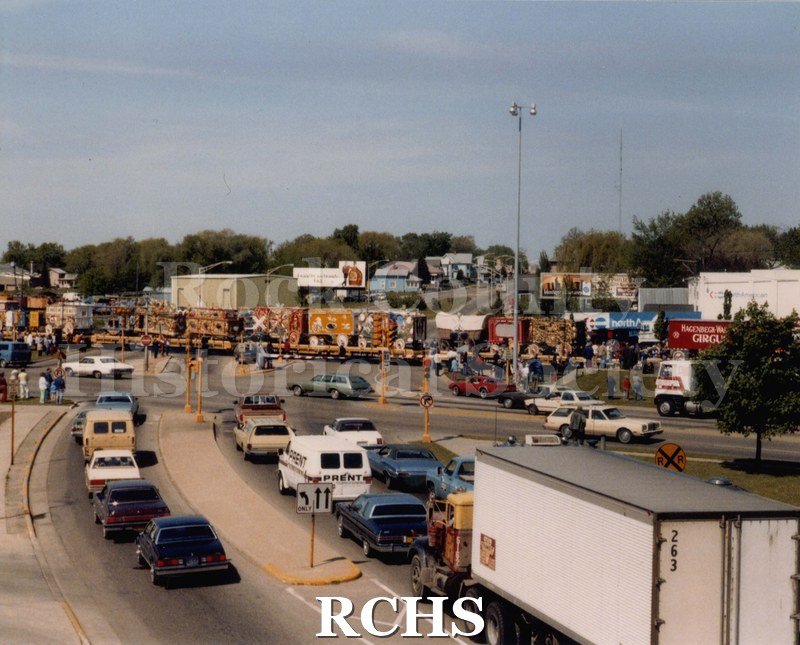 Colored photograph of Baraboo Circus World Parade on train tracks at Five Points intersection, Janesville. Vehicle traffic waiting for crossing.