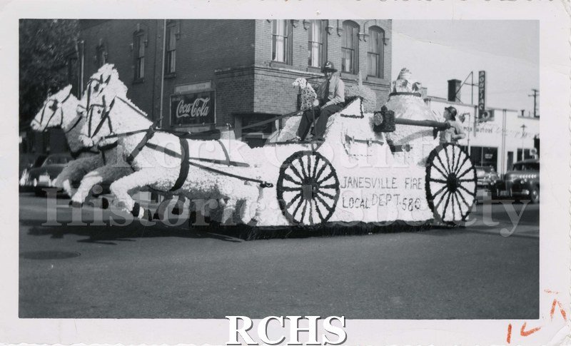 Black and white photograph of the Janesville Fire Dept. parade float. Fire engine with three tissue paper horses with legs above the street.