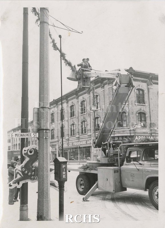 Black and white photograph of decorating for Christmas. Shows city workers downtown on the corner of E. Milwaukee St. hanging Christmas decorations.