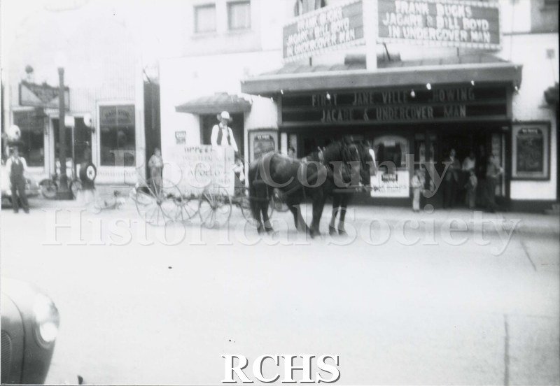 Black and white photograph of the front of Apollo theatre, Janesville, WI. Shows horse and wagon with sign and man standing in wagon.
