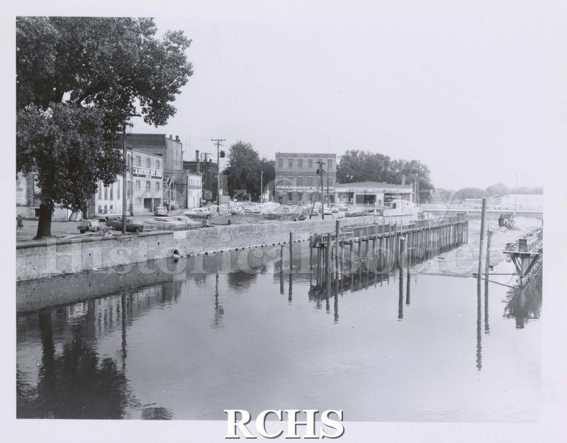 Black and white photograph of Rock River with pilings in river. Chambers & Owens building is in background with many cars parked in back of other buildings.