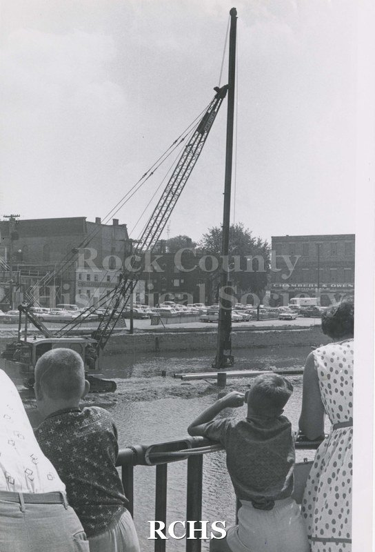 Black and white photograph of construction of parking plaza on Rock River. Shows two boys, man and woman observing the crane setting in a pole.