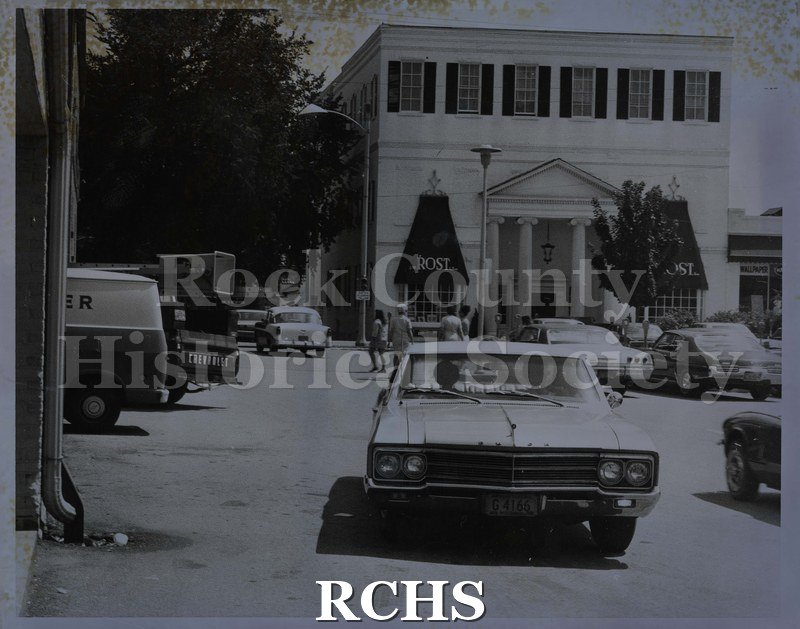 Black and white photograph of cars parked in front of Rost building. Shows several people walking on the street.
