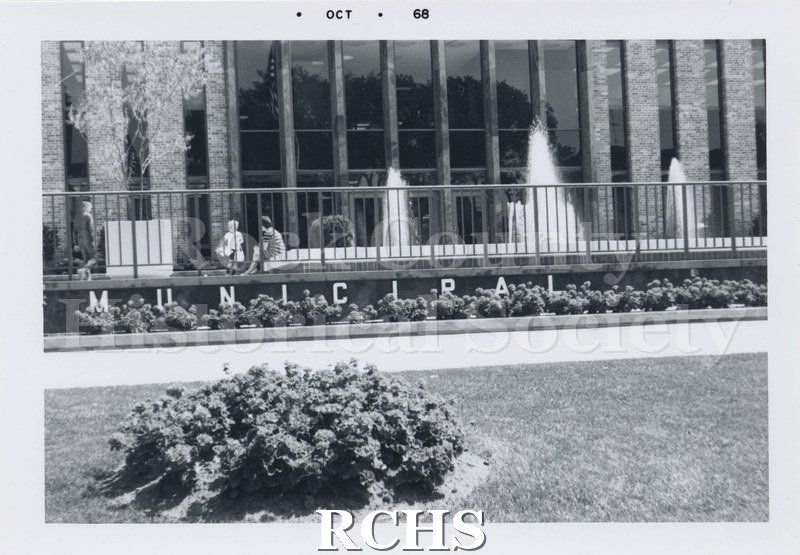 Black and white photograph of Janesville City Hall, showing the front with Municipal spelled out and fountains working. Three people are visible behind the fence.