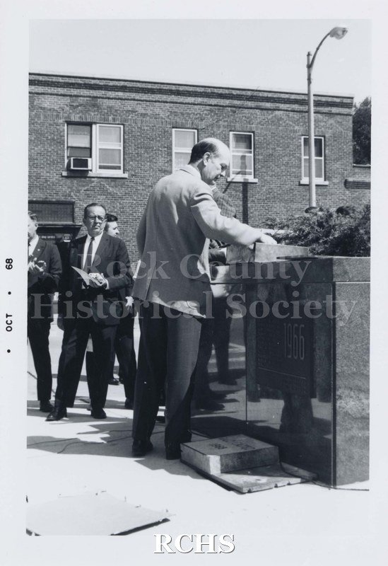 Black and white photograph of an unidentified man standing near a podium. There are other men standing in the background.