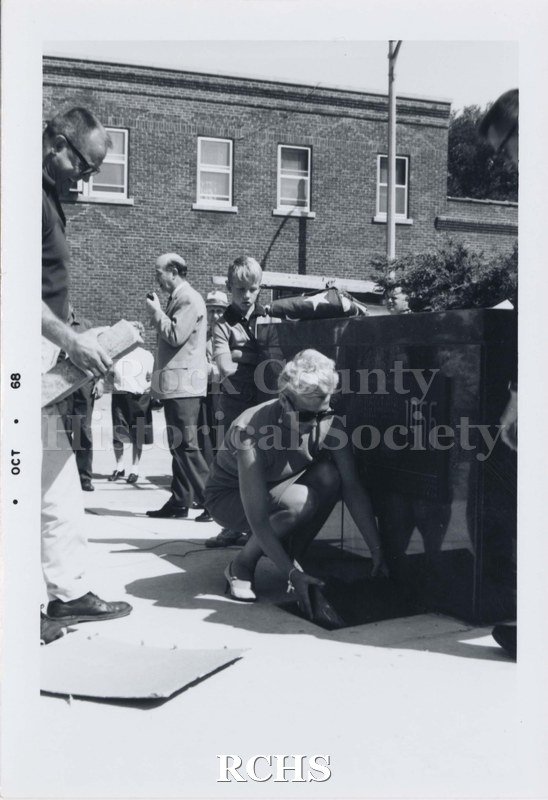 Black and white photograph of an unidentified woman crouched near the 1966 sign. There are two children behind the sign watching.