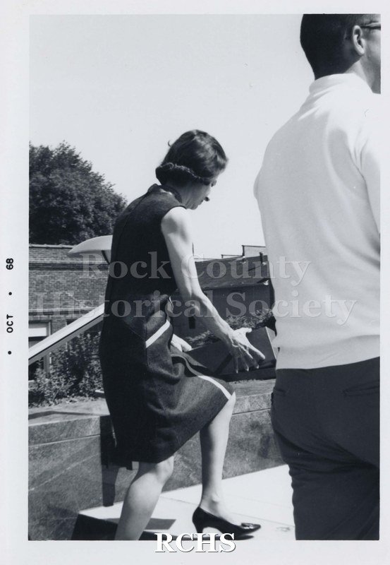 Black and white photograph of unidentified woman walking up a step, carrying a box. She is wearing a dark colored sleeveless dress.