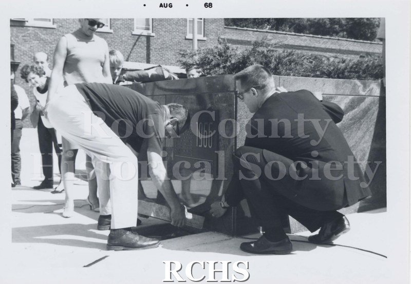 Black and white photograph of two unidentified men standing in front of the plaque 1966. One is bending over and another is kneeling.