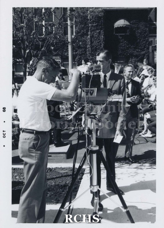 Black and white photograph of an unidentified man taking pictures of man standing behind apparatus. Other people are in the background.