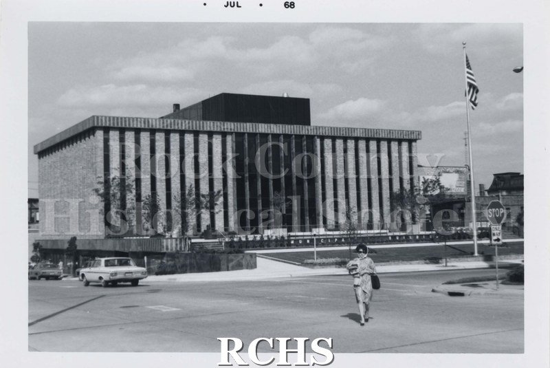Black and white photograph of Janesville City Hall. There is a woman walking across the street; cars parked on the left side of the building and a flagpole with.