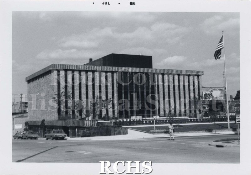 Black and white photograph of Janesville City Hall. There is a woman walking across the street; cars parked on the left side of the building and a flagpole with.