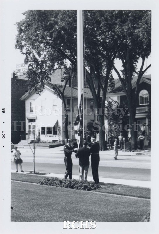 Black and white photograph of flag raising ceremony. Three men are raising the American flag on a flagpole, dressed in uniforms.