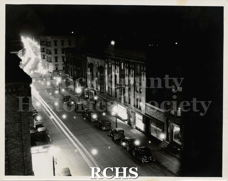 Black and white photograph of East Milwaukee Street, Janesville, WI, taken at night. c. 1925. Shows cars parked on street in front of stores.