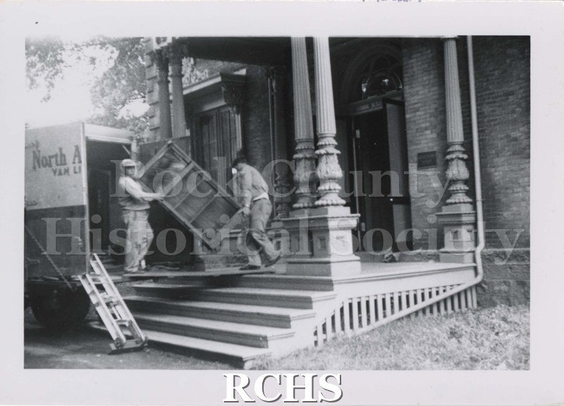 Black and white photograph of two men carrying a bookshelf into the Tallman House. They are standing on the front porch and a van is parked alongside.
