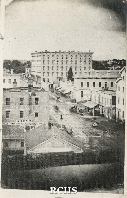 Black and white photograph of Hyatt House in Janesville, WI. Shows buildings beside and in front of hotel, including Addy's Janesville Clothing House.