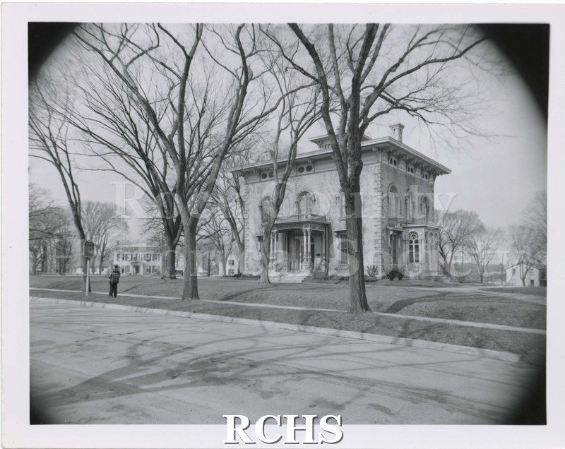 Black and white photograph of the Tallman House taken from road. Shows leafless trees, no fence, and man walking on the sidewalk.