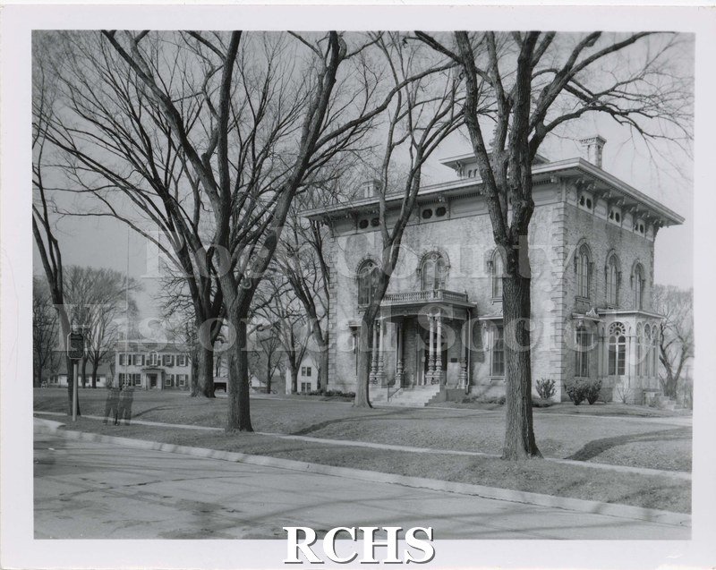 Black and white photograph of the Tallman House taken from road. Shows house from front and east side with trees in front. Another building is visible in background.