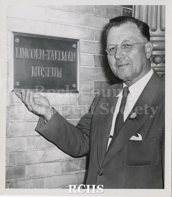 Black and white photograph of John Gross pointing to sign "Lincoln-Tallman Museum".