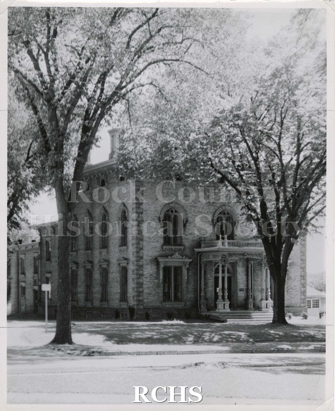 Black and white photograph of the Tallman House taken from road. It shows front and west side of building with two trees just leafing out.