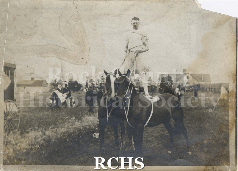 Black and white photograph of a man standing on two black horses wearing a dress. A possible church in the background. Circus wagon (?) in view. Building behind.