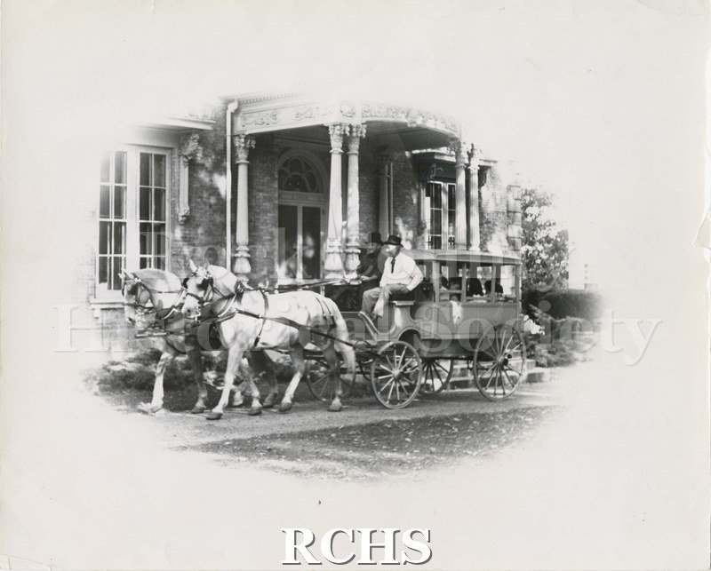 Black and white photograph of Janesville Carriage Work Opera Wagon. Shows a two-horse drawn carriage with two people on the carriage seat parked in front of the.