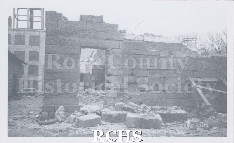 Black and white photograph of the demolition of Rock County Jail. Shows some bricks still standing with other bricks on ground in front of building.