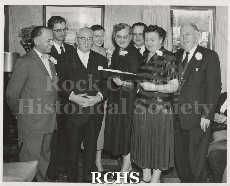 Black and white photograph of the Footville Clinic Dedication. Eight people are standing in a room. The two women are holding a book at which they are looking.