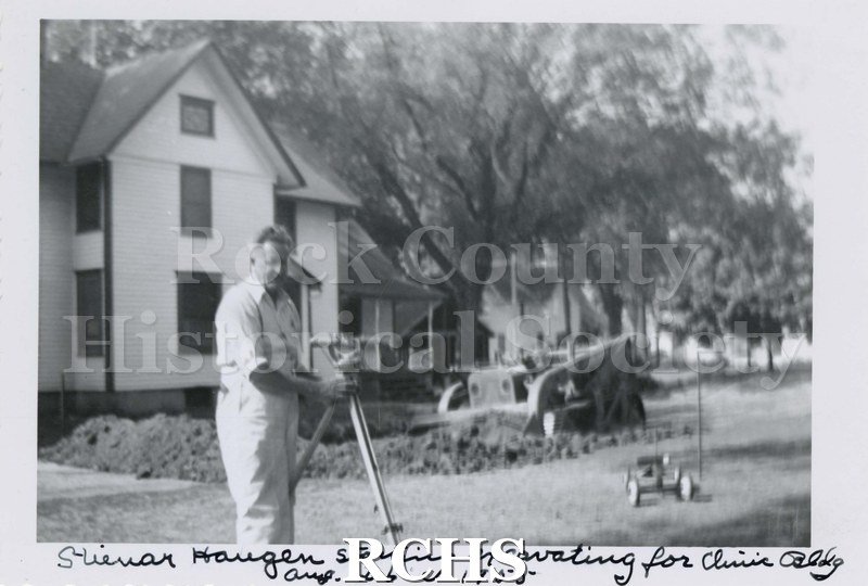 Black and white photograph of Steinar Hauge standing behind a surveyor's device. There is a bulldozer in the background in front of a two story house.
