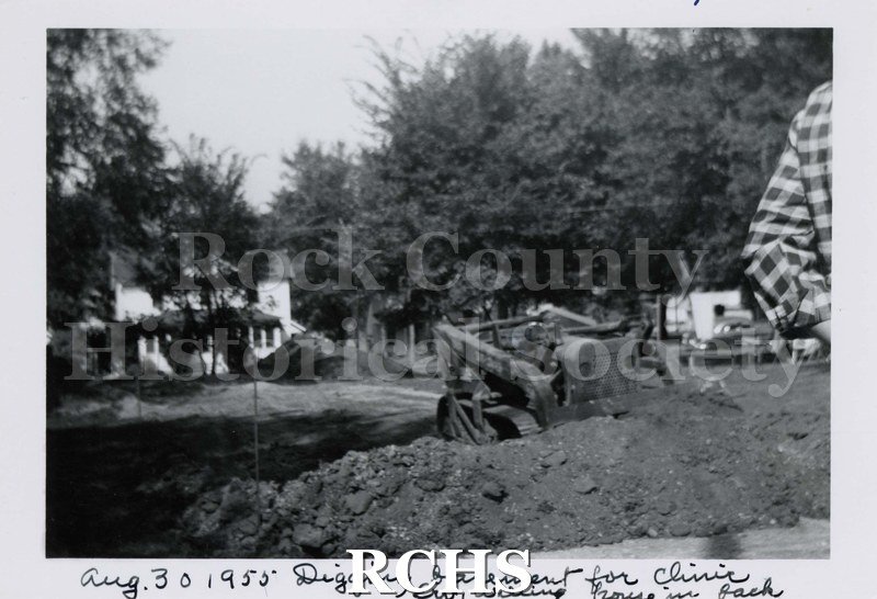 Black and white photograph of a bulldozer excavating a basement. There is a pile of dirt in front of the machine and a man's arm on the right side of the photo.