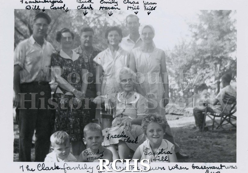 Black and white photograph of the Clark Family. Shows 10 men, women and children with another man sitting in a chair on the right side.