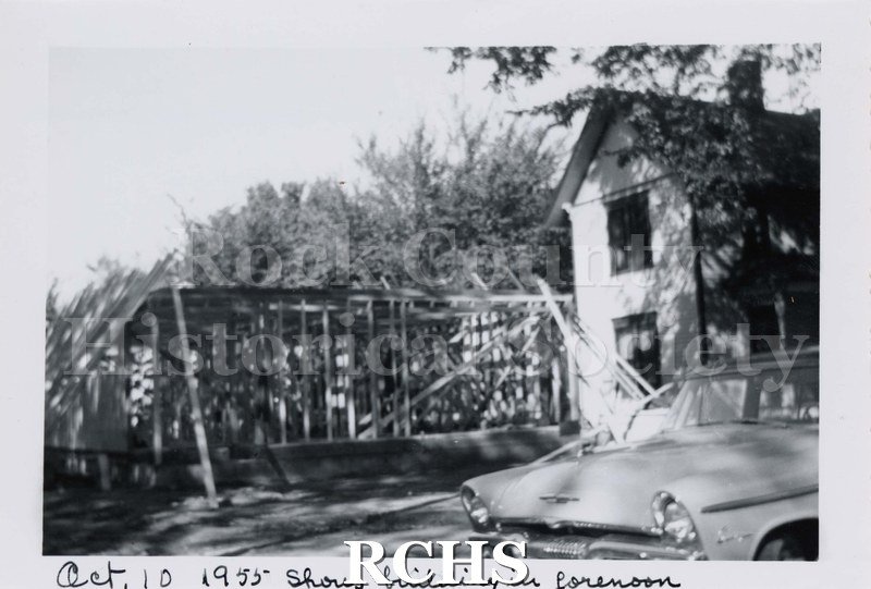Black and white photograph of the Footville Clinic. Shows a partial building beside a two story house. There is a car in the foreground.
