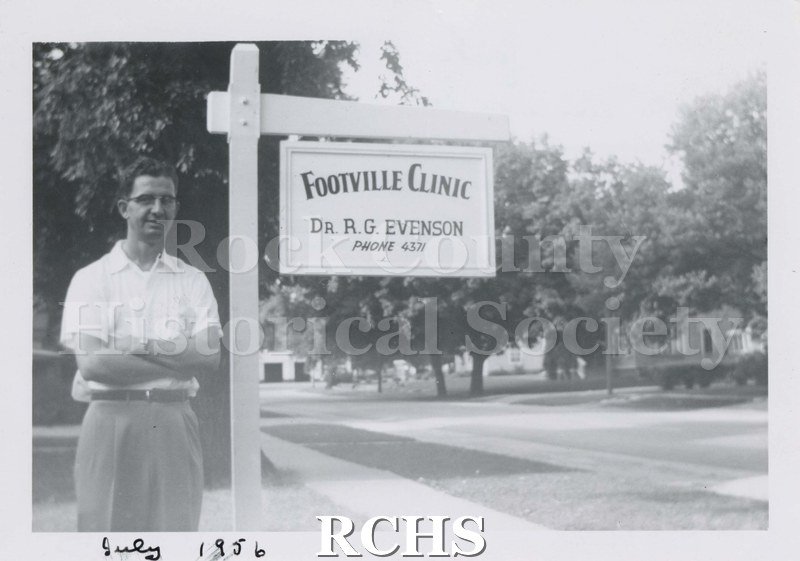 Black and white photograph of Dr. R. G. Evenson standing beside a sign which reads: "Footville Clinic Dr. R. G. Evenson Phone 4371.".