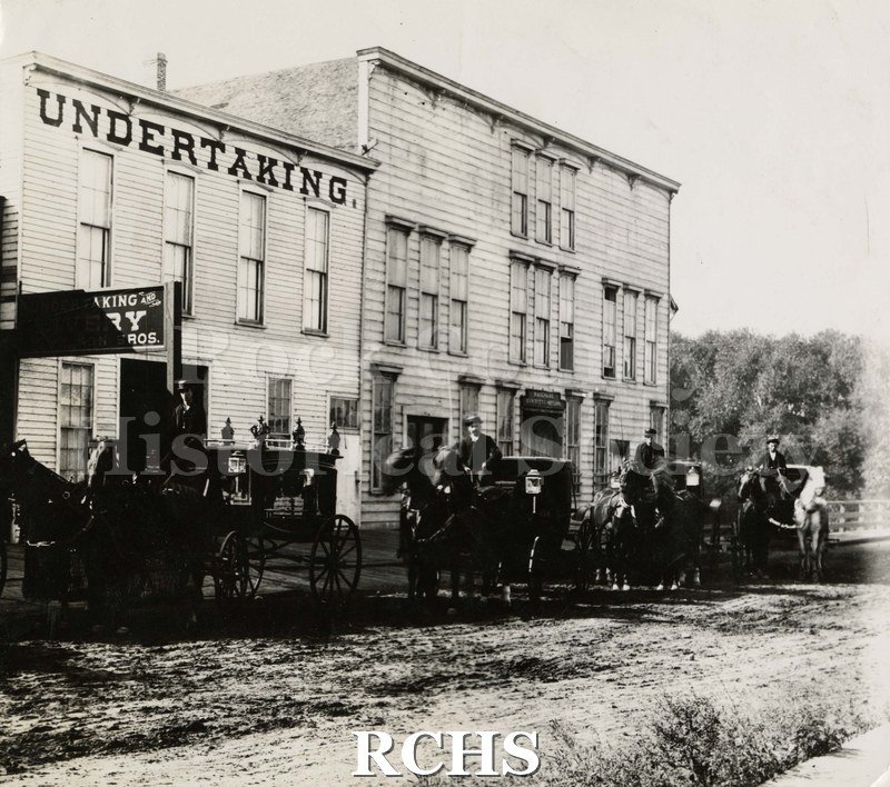 Black and white photograph of early Janesville buildings. One building has "Undertaking" on top and another sign partially blocked reads "...king".