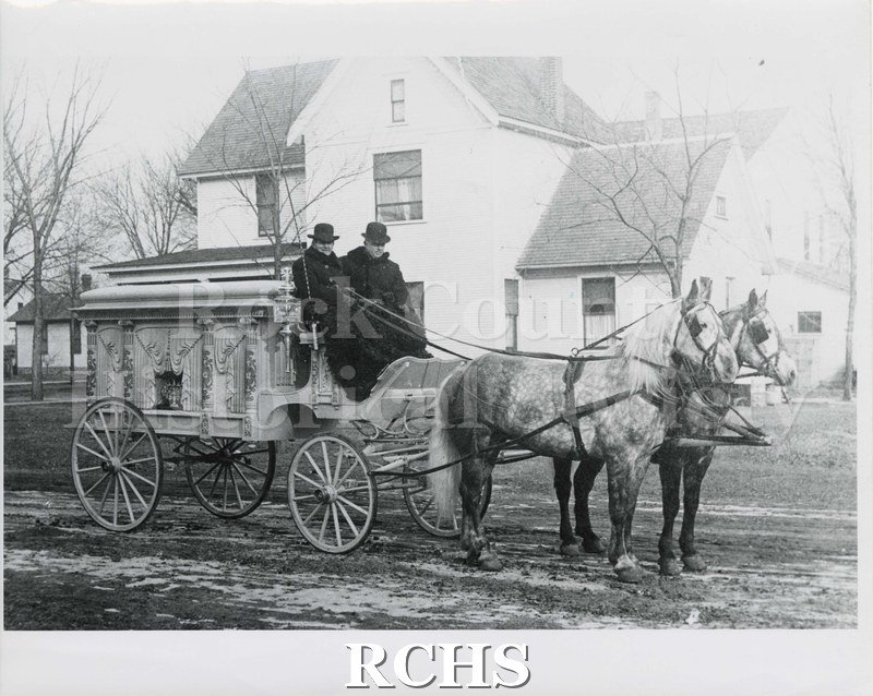 Black and white photograph of a Janesville funeral hearse pulled by two dappled gray horses. There are two men seated on the hearse.