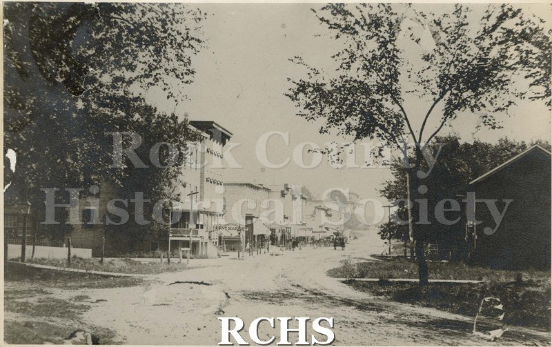Black and white photograph of Edgerton, WI. 1886. Shows buildings on left side with a carriage on the street. There is a building with a tree on the right side.