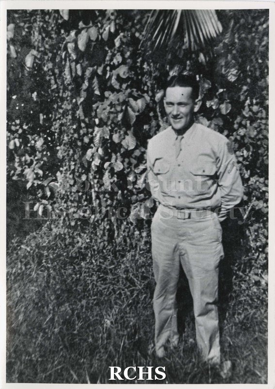 Black and white photograph of Glen Swinbank, a soldier, standing in front of vegetation. He is wearing a uniform and has his hands behind his back.