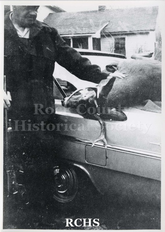 Black and white photograph of Harry Johnson. Shows a man standing beside a car with a dead deer on car.